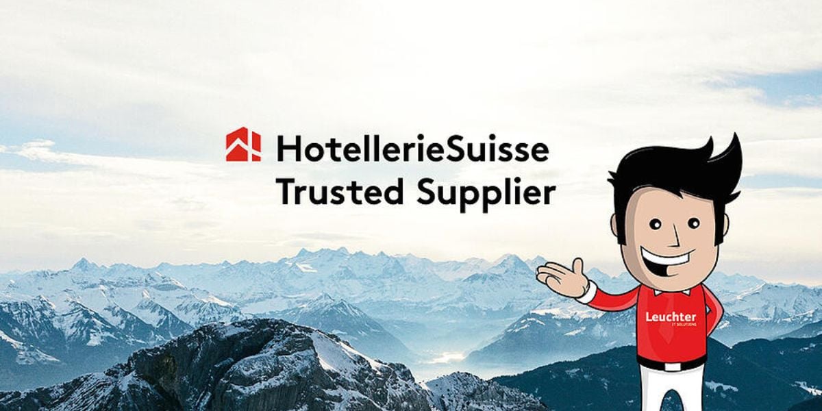 HotellerieSuisse Trusted Supplier – Leuchter IT-Maskottchen vor Schweizer Alpenpanorama mit Auszeichnung für Hotelpartner
