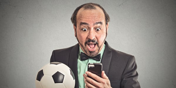 Mann im Anzug mit Fussball in der Hand schaut begeistert auf Smartphone