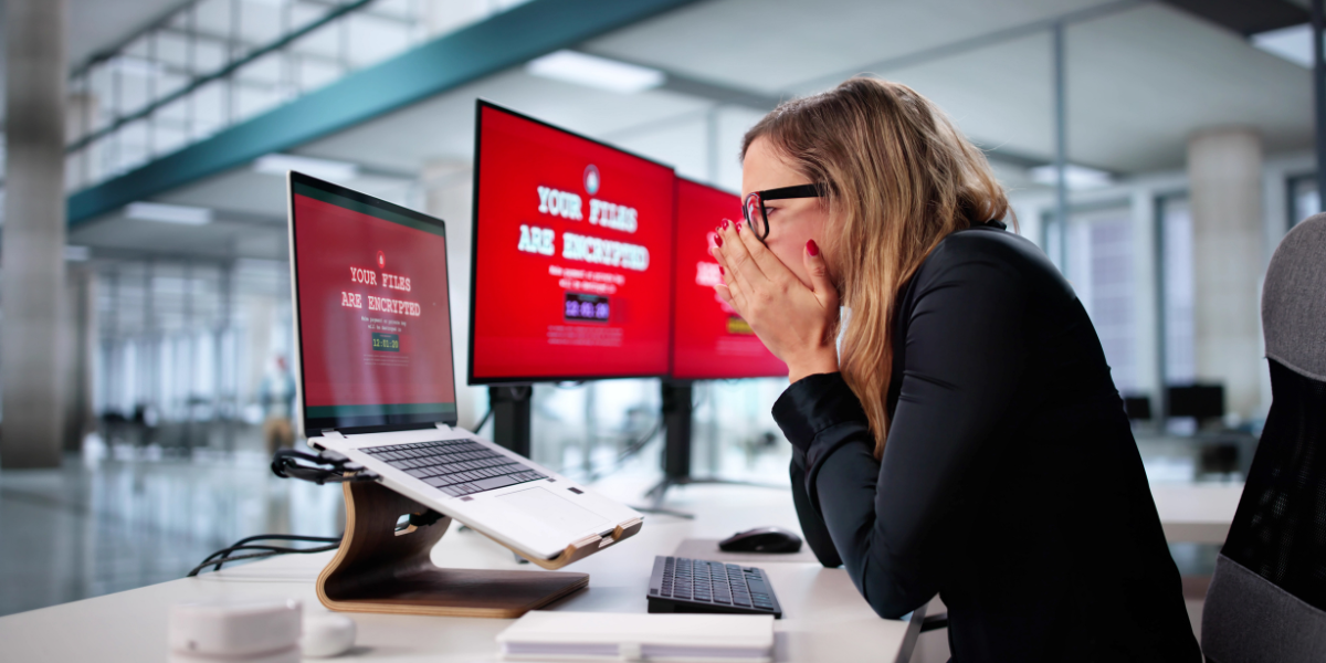 Eine Frau sitzt besorgt an einem Schreibtisch und blickt auf drei Computerbildschirme mit der Meldung „Ihre Dateien sind verschlüsselt