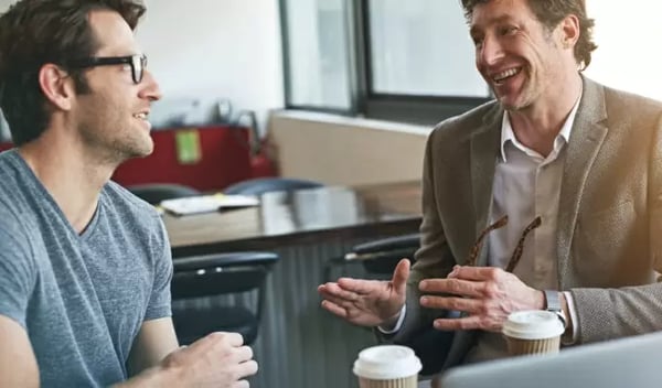 Kommunikation im Büro: Zwei männliche Kollegen unterhalten sich entspannt bei Kaffee. Der ältere Mann gestikuliert lächelnd, während der jüngere interessiert zuhört.
