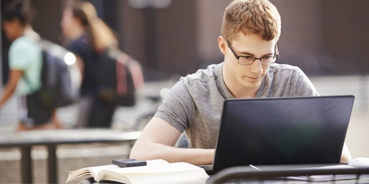 Student mit Laptop beim Lernen im Freien – junger Mann mit Brille arbeitet am Notebook neben Buch und Smartphone