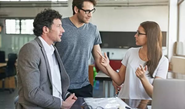 Organisation im Büro: Drei Kollegen in einer modernen Arbeitsumgebung diskutieren intensiv ein Projekt. Eine Frau mit Brille gestikuliert erklärend, während zwei Männer aufmerksam zuhören.