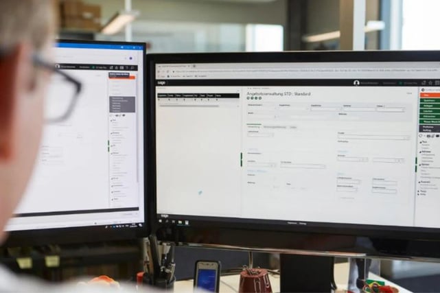 Blick über die Schulter einer Fachkraft, die 'Sage 100' für Rechnungswesen auf zwei Computermonitoren in einem Schweizer Büro verwendet.