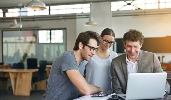 Zusammenarbeit im Büro: Drei Kollegen schauen gemeinsam auf einen Laptop-Bildschirm und lachen. Die lockere Atmosphäre zeigt produktives Teamwork in einem modernen Arbeitsumfeld.